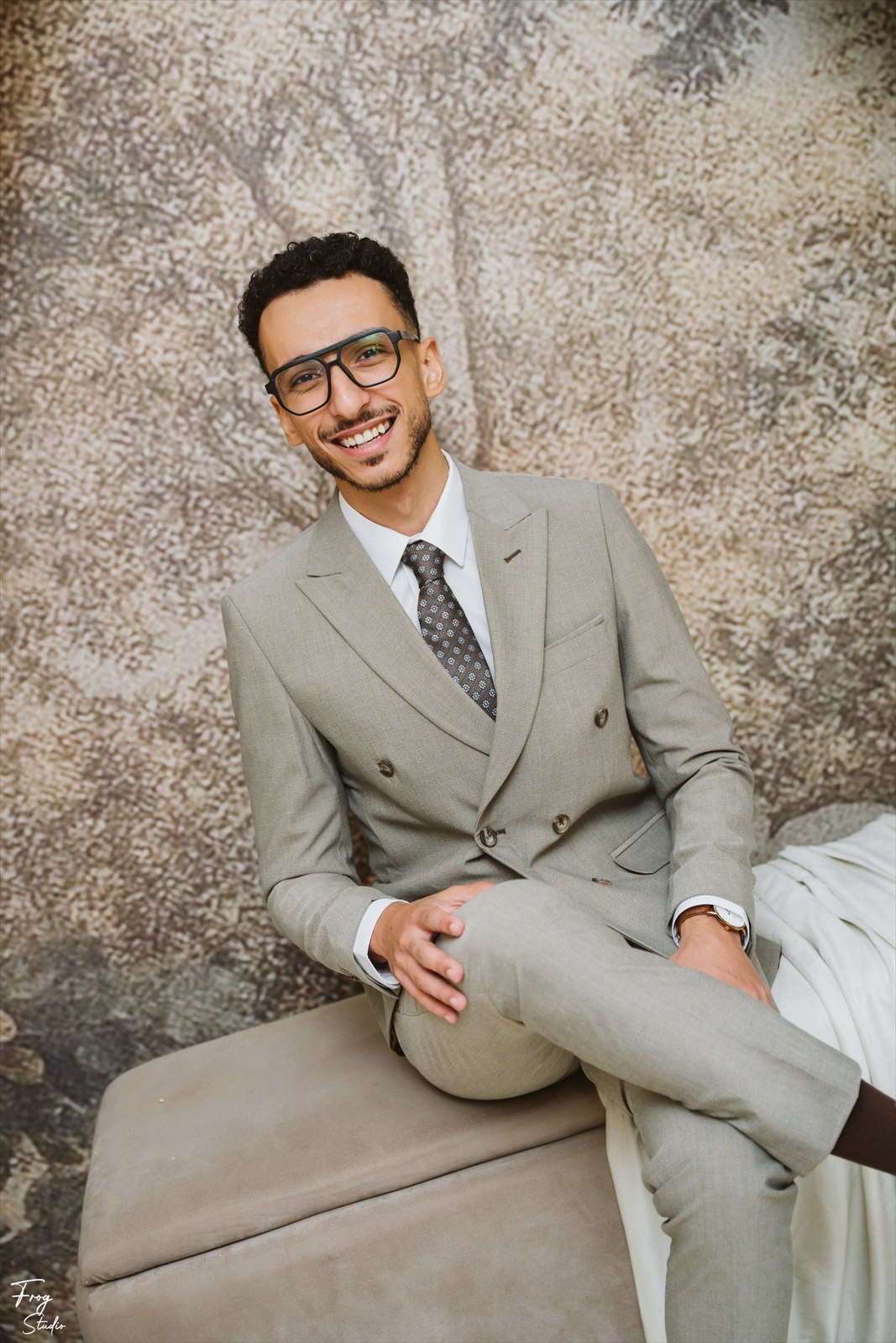 Mohamed seated on an ottoman in his grey double-breasted suit, patterned tie and glasses, smiling
