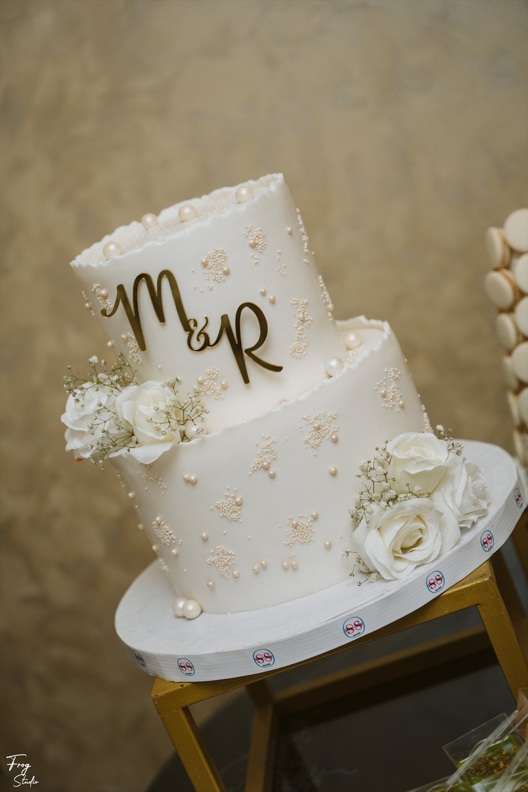 Two-tier white engagement cake with pearl detailing, gold M&R monogram and white roses