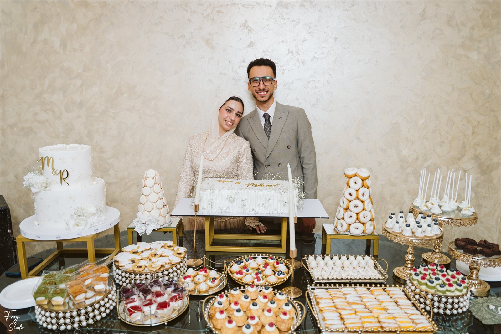 Reem and Mohamed standing together behind their engagement sweet table, framed by the monogram cake, macaron tower and gold platters